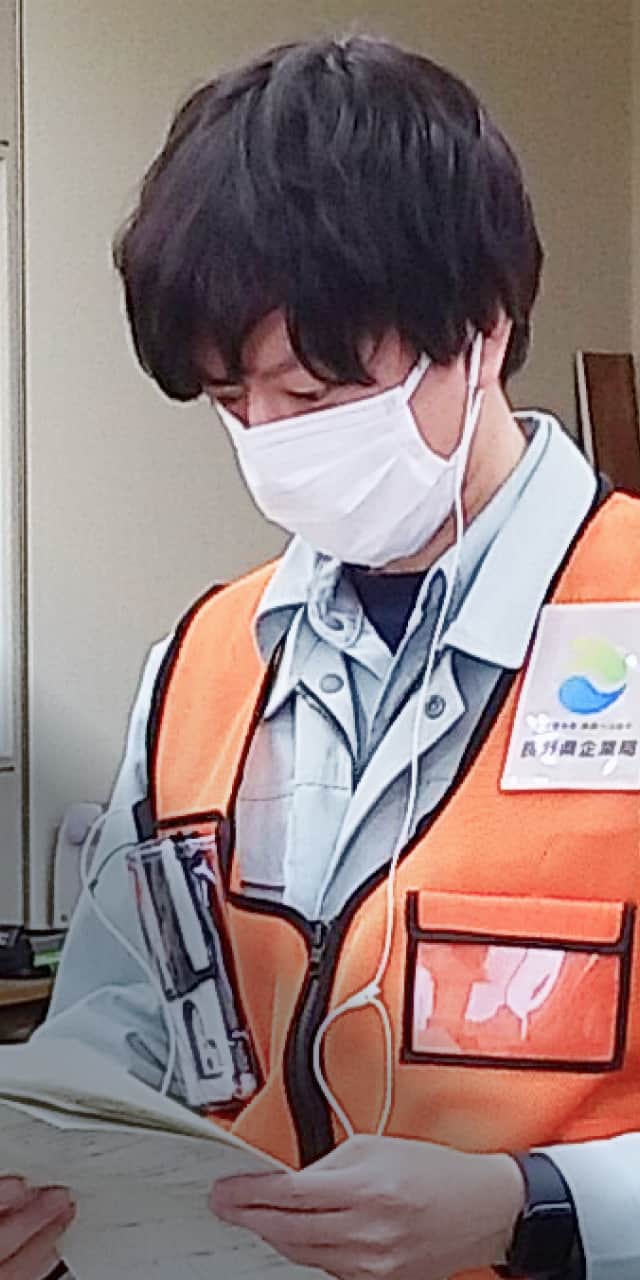 オレンジの作業服を着た男性が、マスクを装着しながらノートを見つめている室内の様子。