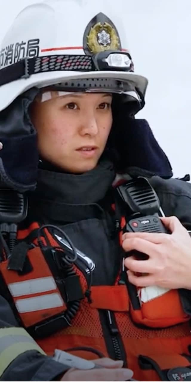 雪の中で消防士の女性が通信機器を持ち、真剣な表情で前方を見つめている。