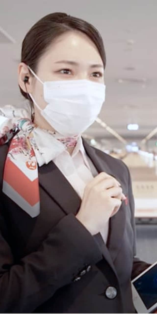 空港のカウンターで、マスクをつけた女性スタッフが手にタブレットを持ち、微笑みながら話している。