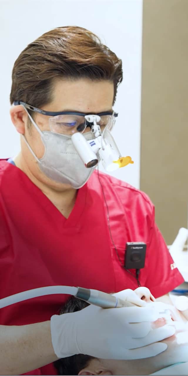 赤い医療ユニフォームを着た医師が患者の口腔内を治療している場面で、顕微鏡が装着された眼鏡を使用している。