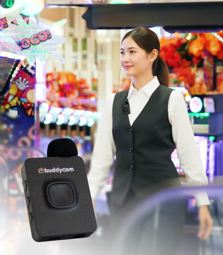 カジュアルな服装の女性がカジノ風のゲームエリアで笑顔で歩いており、胸元にはbuddycomのマイクがある。