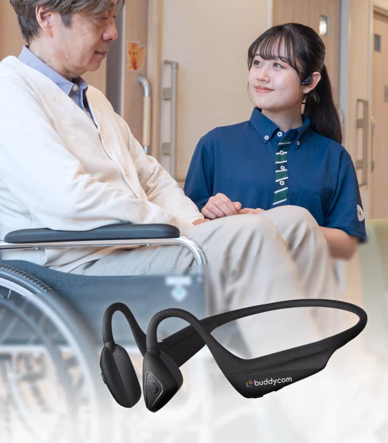 車椅子に座る男性と、彼に話しかける女性がいる室内で、buddycomのデバイスが手前に置かれている。